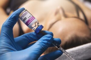 A close-up of a gloved hand holding a vial of botulinum toxin and a medical syringe, with a patient's face in soft focus in the background, illustrating a professional BOTOX® treatment setting.