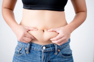 Woman in black crop top and jeans pinching belly fat around her midsection against a light gray background