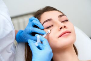 A medical professional wearing blue nitrile gloves carefully administers a dermal filler injection into the tear trough area below the eye of a patient.